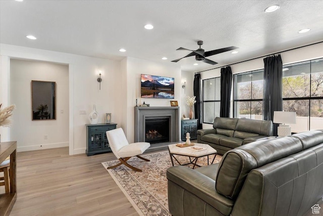 living area featuring light wood-type flooring, baseboards, recessed lighting, a ceiling fan, and a warm lit fireplace