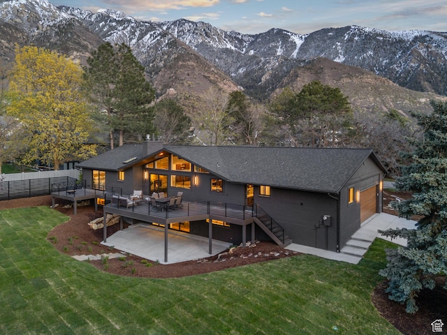 rear view of house with a deck with mountain view, fence, a shingled roof, a yard, and a patio
