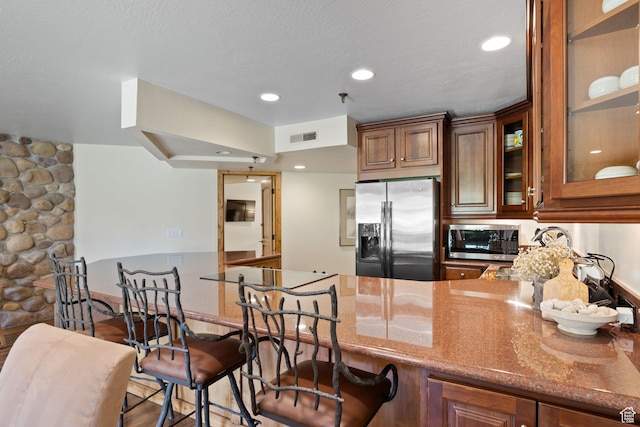 kitchen with brown cabinets, a breakfast bar area, glass insert cabinets, stone counters, and appliances with stainless steel finishes