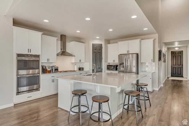 kitchen featuring wall chimney exhaust hood, stainless steel appliances, a sink, a center island with sink, and a kitchen bar