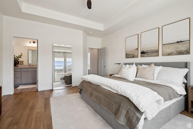bedroom featuring baseboards, wood finished floors, ensuite bathroom, and a raised ceiling
