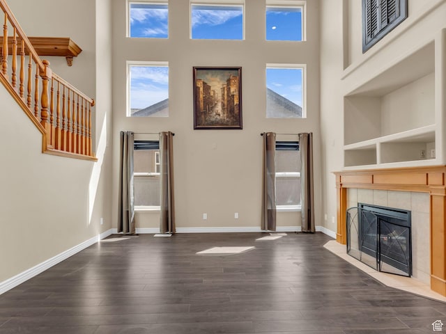 unfurnished living room featuring a high ceiling, dark wood-type flooring, a tiled fireplace, baseboards, and stairway