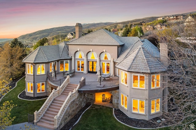 back of property at dusk with stairway, a yard, a deck with mountain view, a chimney, and stucco siding