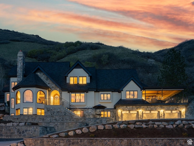 back of property at dusk featuring a balcony, a mountain view, stone siding, a chimney, and stucco siding