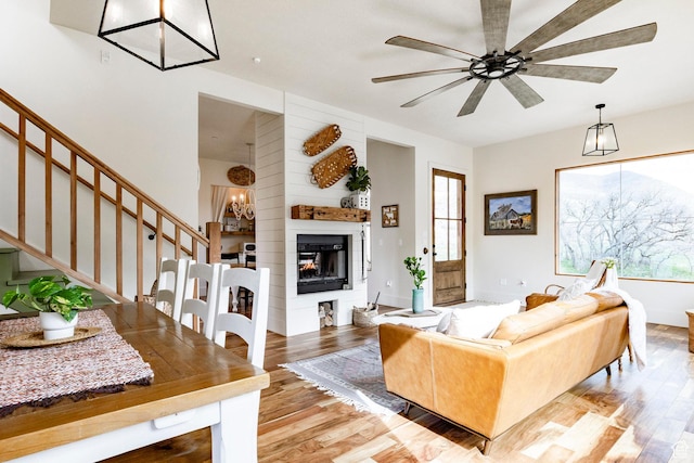 living area featuring stairs, a glass covered fireplace, wood finished floors, and ceiling fan with notable chandelier