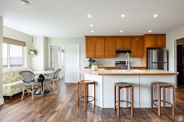 kitchen with brown cabinetry, stainless steel appliances, under cabinet range hood, light countertops, and dark wood-type flooring