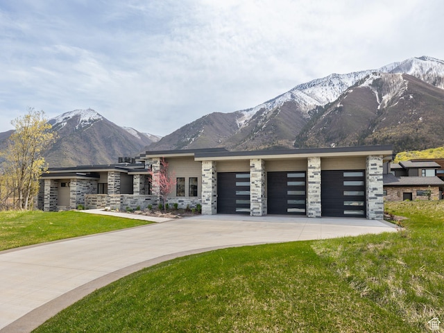 prairie-style home with stone siding, a mountain view, a front yard, concrete driveway, and an attached garage
