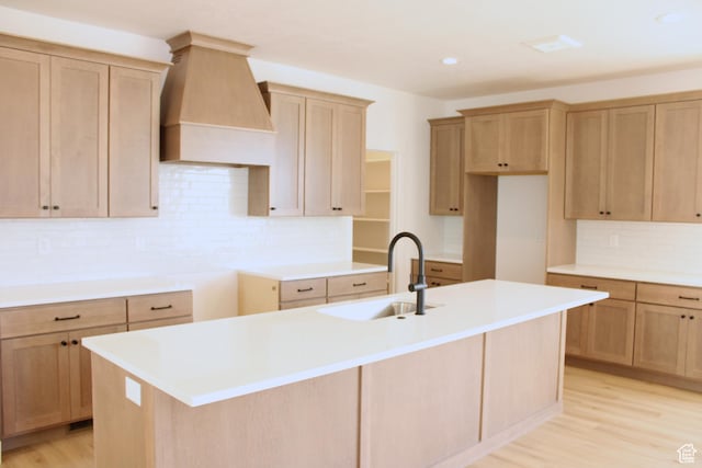 kitchen with a kitchen island with sink, decorative backsplash, light wood-style floors, a sink, and premium range hood