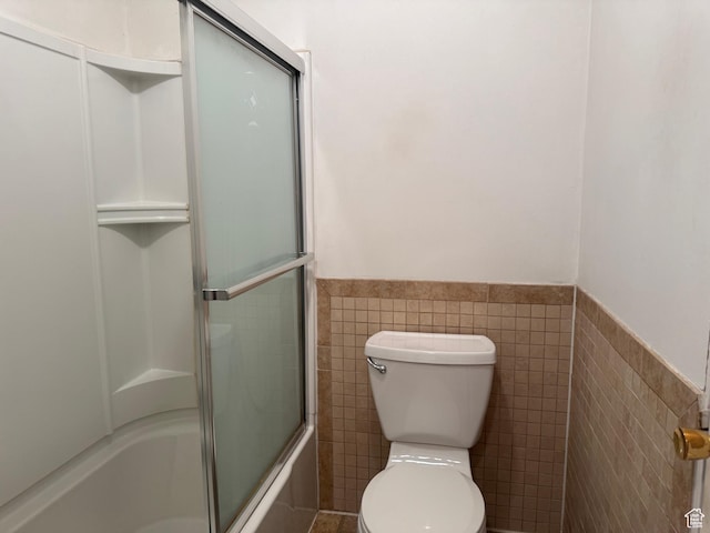 full bath featuring combined bath / shower with glass door, toilet, wainscoting, and tile walls