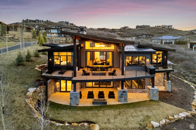 back of house at dusk featuring a standing seam roof, a balcony, a patio, and stone siding