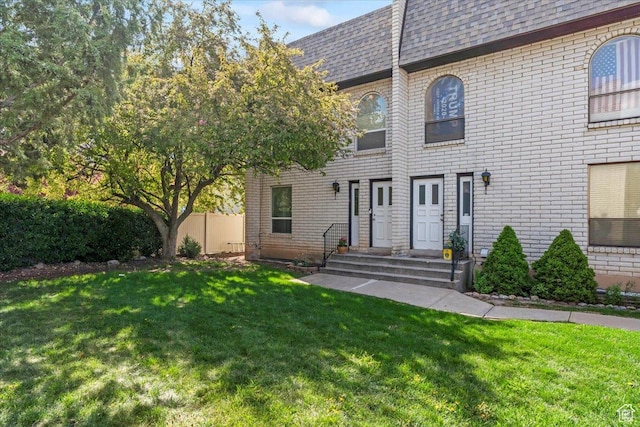 view of front of home featuring brick siding, fence, roof with shingles, and a front lawn