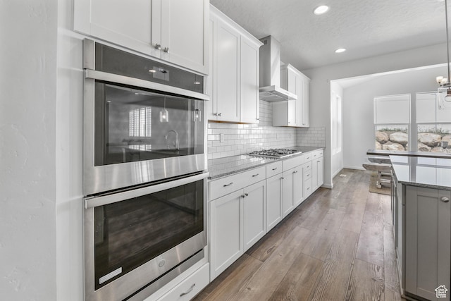 728 W Wild Hyacinth Dr, Saratoga Springs, UT 84045 - kitchen featuring wall chimney range hood, light stone countertops, stainless steel appliances, white cabinetry, and tasteful backsplash