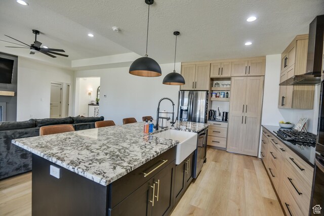 kitchen with light brown cabinetry, wall chimney range hood, stainless steel fridge, a sink, and light wood finished floors