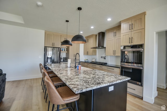 kitchen featuring appliances with stainless steel finishes, light brown cabinetry, a sink, baseboards, and wall chimney exhaust hood