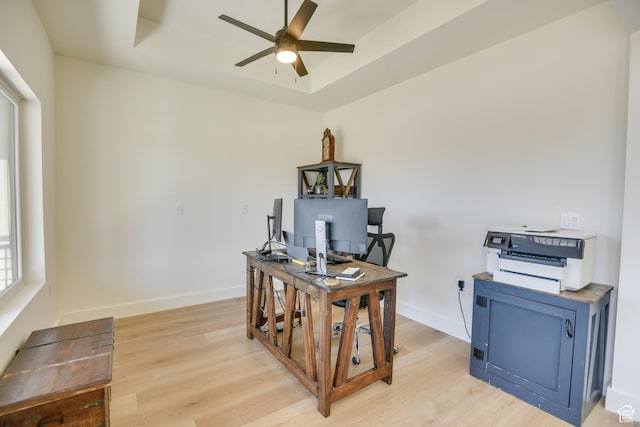 office featuring a ceiling fan, a tray ceiling, baseboards, and light wood-type flooring