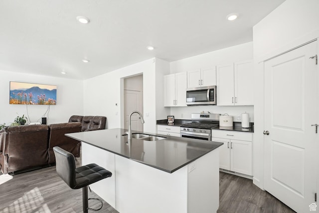 kitchen with dark countertops, an island with sink, dark wood-type flooring, a sink, and appliances with stainless steel finishes