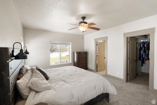 bedroom featuring light colored carpet, a walk in closet, a textured ceiling, a ceiling fan, and baseboards