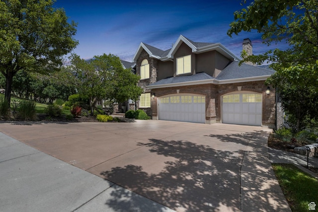 view of front of house featuring a shingled roof, stone siding, a garage, a chimney, and driveway