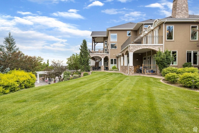 rear view of house with a lawn, a balcony, a patio, stone siding, and stucco siding