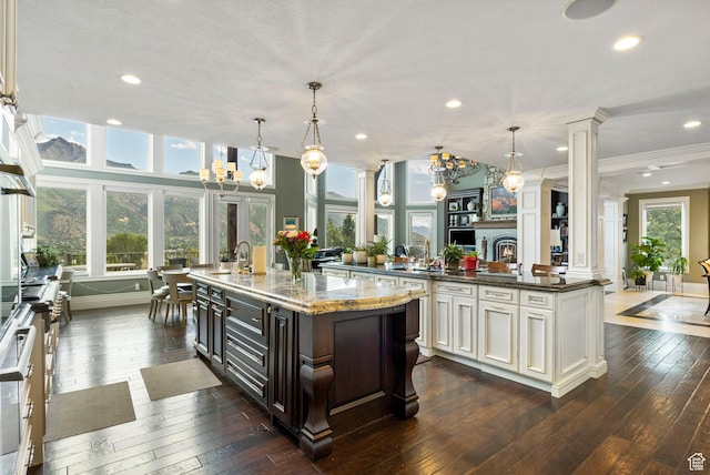 kitchen with decorative columns, crown molding, a large island with sink, an inviting chandelier, and dark wood-type flooring