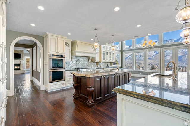 kitchen featuring stainless steel double oven, a center island with sink, a sink, baseboards, and custom exhaust hood