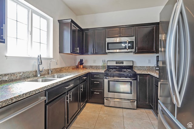 kitchen featuring dark brown cabinetry, appliances with stainless steel finishes, a sink, light tile patterned floors, and light stone counters