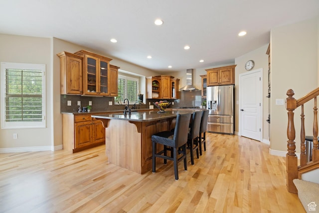 kitchen featuring a breakfast bar area, light wood-style floors, wall chimney exhaust hood, stainless steel refrigerator with ice dispenser, and a center island with sink