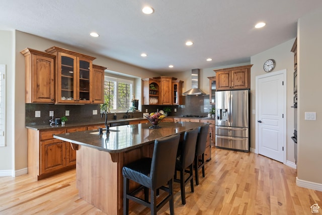 kitchen featuring a sink, brown cabinets, wall chimney exhaust hood, an island with sink, and stainless steel fridge