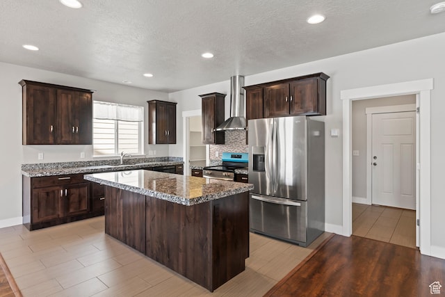 kitchen with a kitchen island, dark brown cabinets, wall chimney exhaust hood, light wood-style flooring, and appliances with stainless steel finishes