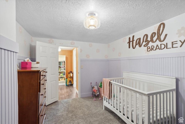 carpeted bedroom featuring a nursery area, a textured ceiling, and wainscoting