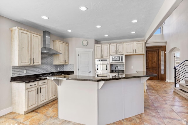 kitchen with decorative backsplash, baseboards, a center island with sink, wall chimney range hood, and stainless steel appliances