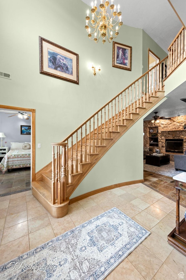 staircase with visible vents, ceiling fan with notable chandelier, a high ceiling, and a stone fireplace