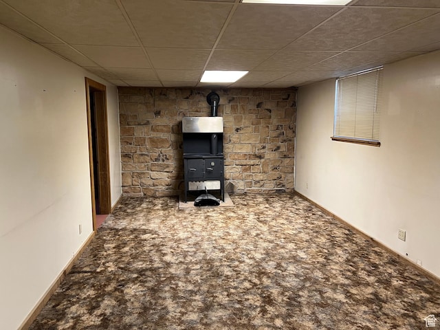 finished basement featuring a drop ceiling, carpet, baseboards, and a wood stove