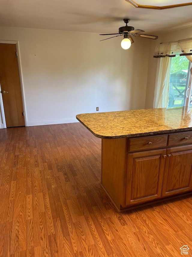 kitchen featuring wood finished floors, a ceiling fan, brown cabinets, and light stone countertops
