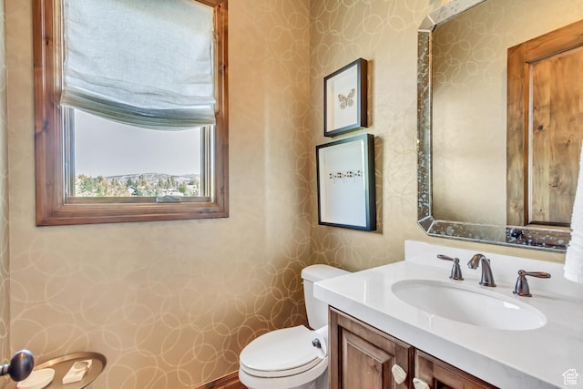 half bathroom with vanity, toilet, and wallpapered walls