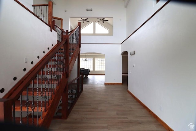 staircase with wood finished floors, a towering ceiling, arched walkways, a ceiling fan, and baseboards