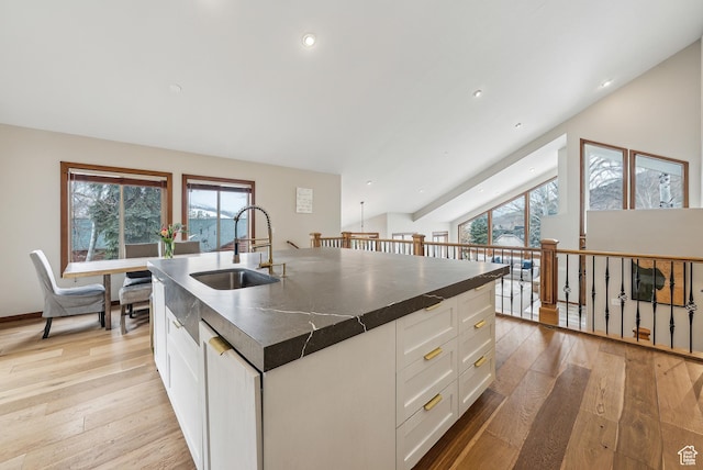 kitchen with light wood-style flooring, dark countertops, a sink, a center island with sink, and white cabinets