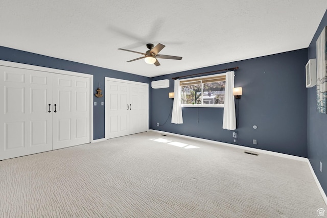 unfurnished bedroom with a wall unit AC, two closets, baseboards, carpet flooring, and a ceiling fan