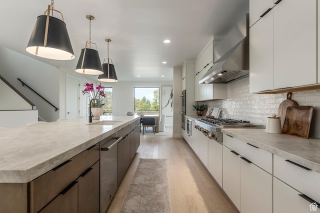 kitchen with light wood-style flooring, stainless steel appliances, a sink, wall chimney exhaust hood, and light countertops
