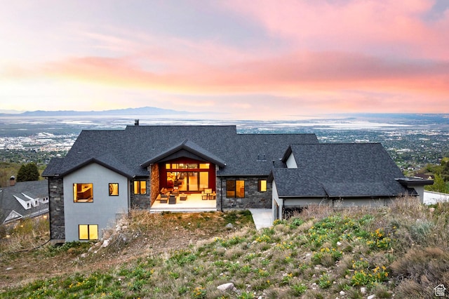 back of property at dusk with roof with shingles, stone siding, and a mountain view