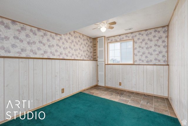 empty room featuring a ceiling fan, wainscoting, baseboards, wallpapered walls, and wooden walls