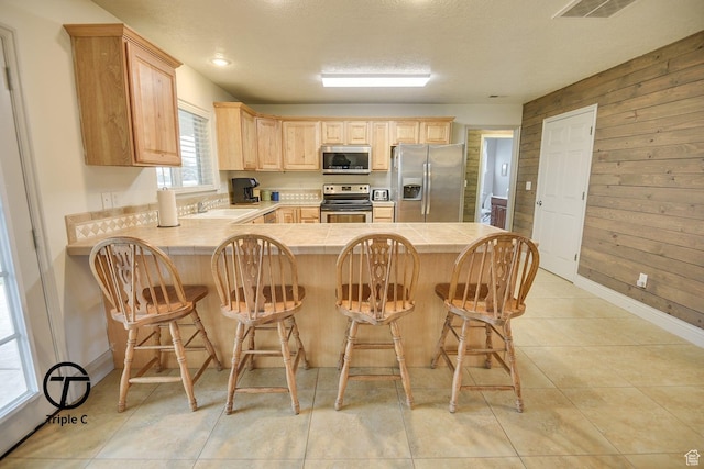 kitchen featuring appliances with stainless steel finishes, light brown cabinetry, wood walls, tile countertops, and a peninsula
