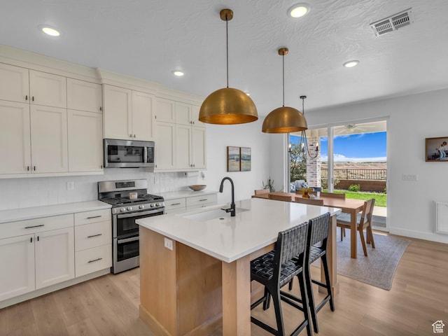 kitchen featuring stainless steel appliances, a sink, backsplash, light wood-style floors, and a center island with sink