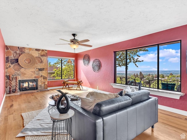 living room featuring a textured ceiling, ceiling fan, wood finished floors, a stone fireplace, and baseboards