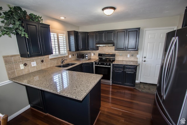 kitchen featuring appliances with stainless steel finishes, a sink, under cabinet range hood, a peninsula, and dark cabinets