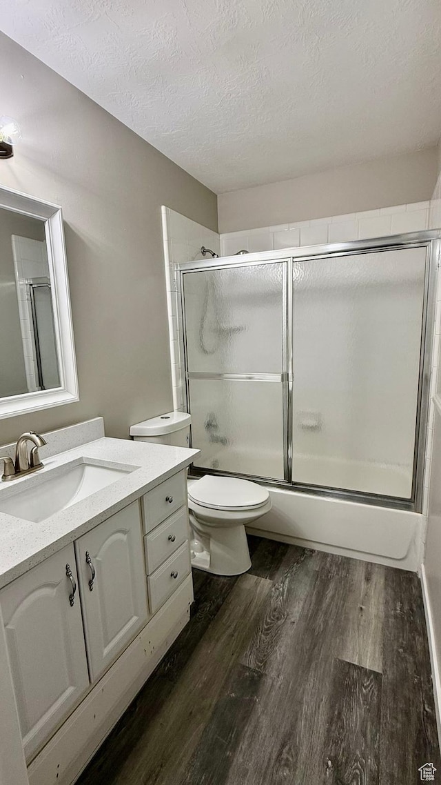 bathroom with a textured ceiling, wood finished floors, toilet, bath / shower combo with glass door, and vanity