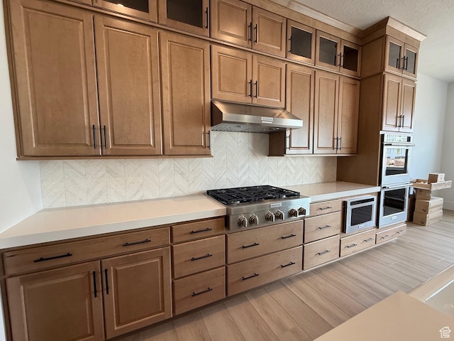 kitchen featuring under cabinet range hood, stainless steel appliances, light wood-style floors, decorative backsplash, and a textured ceiling