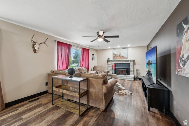 living room featuring ceiling fan, a stone fireplace, dark wood-type flooring, a textured ceiling, and baseboards