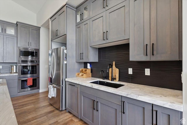 kitchen featuring appliances with stainless steel finishes, a sink, gray cabinets, tasteful backsplash, and glass insert cabinets