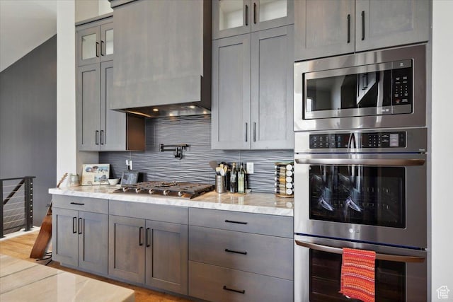 kitchen featuring appliances with stainless steel finishes, backsplash, custom exhaust hood, glass insert cabinets, and light stone counters